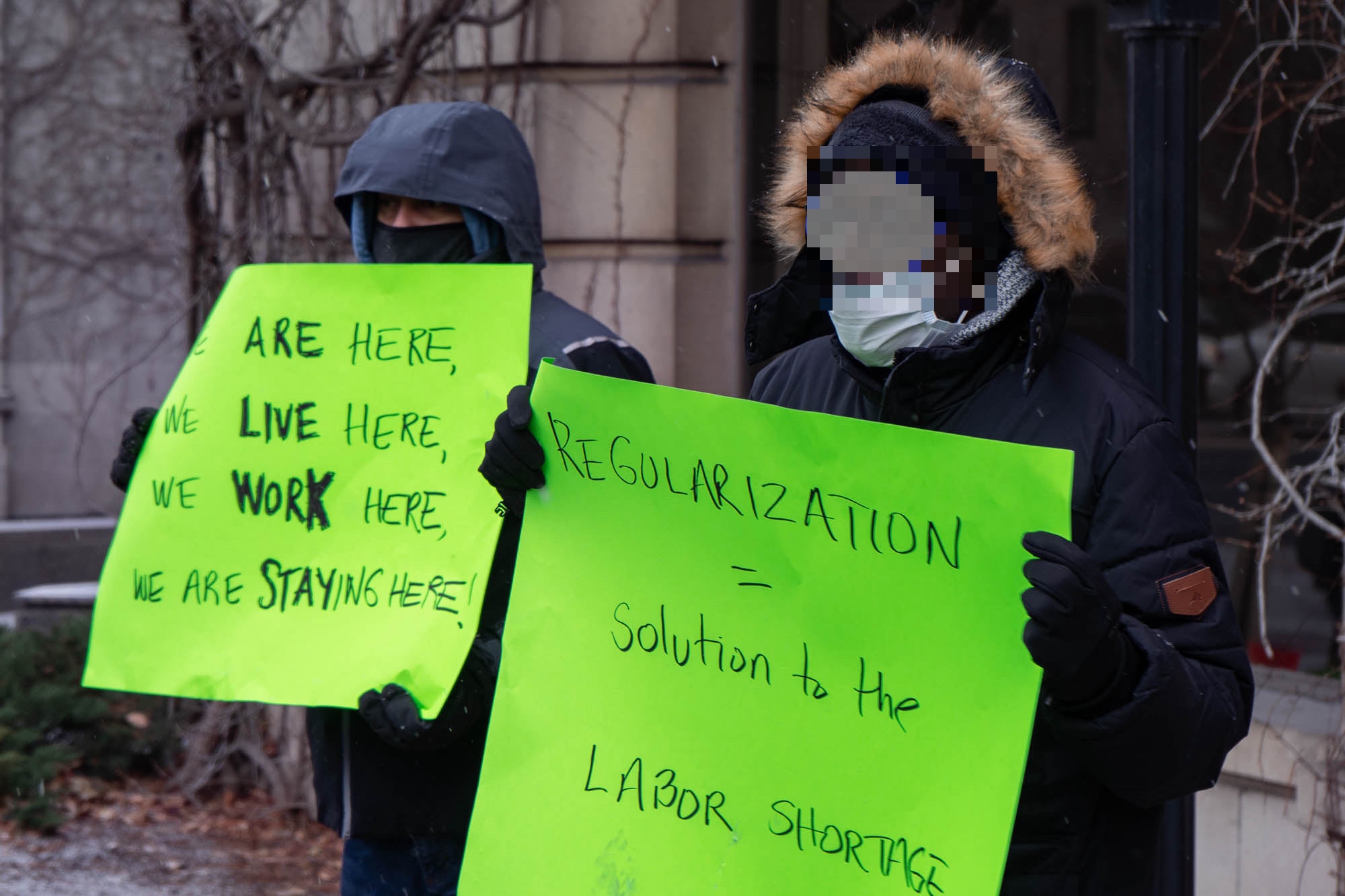 Two racialized people hold up signs outside in winter. One is light green with black text reading "Regularisation = solution to the labour shortage." Photo from a Solidarity Across Borders event in December 2021 in Montreal, Undocumented migrants have a solution to the labor shortage: Status for all. Photo: Solidarity Across Borders.