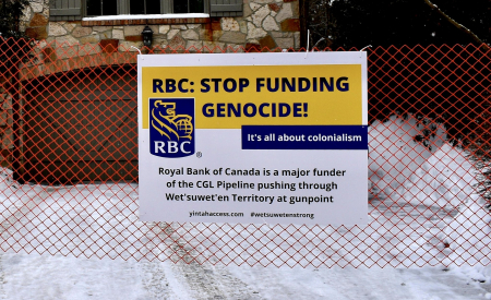 A sign is placed on orange construction mesh in front Doug Guzman's house, reading "RBC: STOP FUNDING GENOCIDE!" Photo credit: World BEYOND War Canada